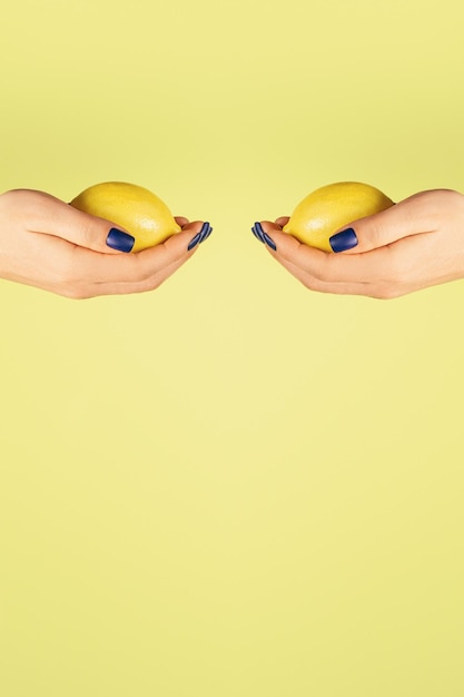 Dos manos femeninas abiertas con uñas azules sosteniendo limones frescos uno frente al otro aislado en amarillo. Vertical. Copia espacio