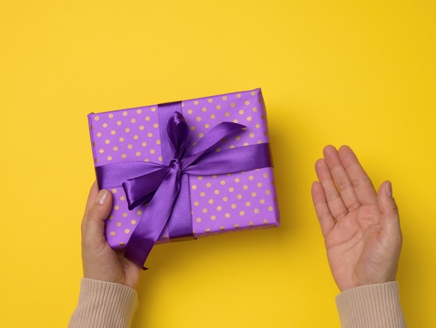 Dos manos femeninas sostienen una caja de regalo de color púrpura sobre un fondo amarillo, concepto de feliz cumpleaños, vista superior