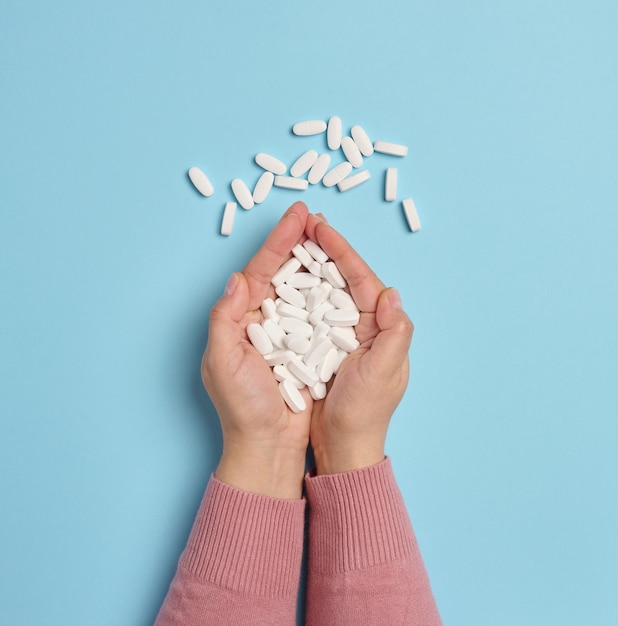 Dos manos femeninas sostienen pastillas ovaladas blancas en un fondo azul vista superior