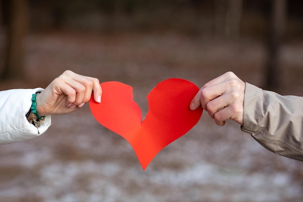 Dos manos, masculina y femenina, rasgan el símbolo del corazón rojo de los amantes contra el fondo del bosque invernal. Concepto de romper la relación. El amor ha terminado. Resentimiento y separación. Negativo.