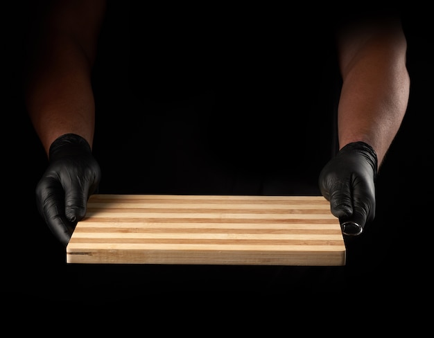 Dos manos masculinas en guantes de látex negro sostienen una nueva tabla de cortar de madera marrón vacía, fondo negro