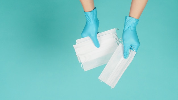 Dos manos sosteniendo máscaras faciales usan guantes médicos azules sobre fondo verde menta o azul Tiffany