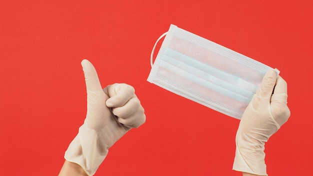 Dos manos usan guantes de látex o guantes médicos blancos y mascarilla sobre fondo rojo.