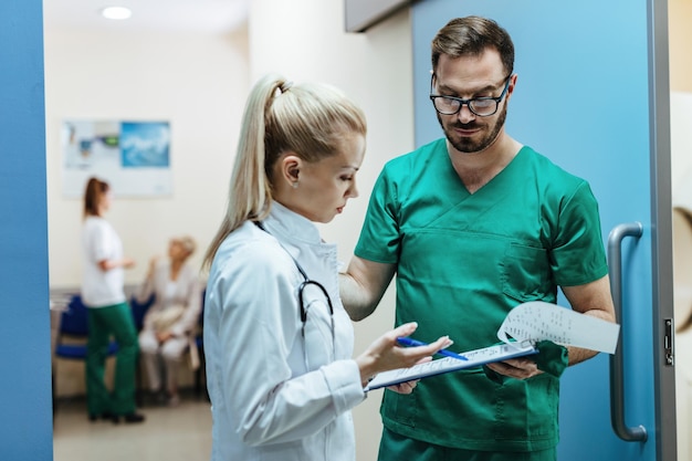 Dos médicos analizando informes médicos mientras están de pie en el vestíbulo de la clínica El foco está en un cirujano