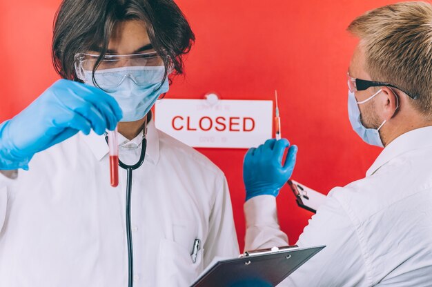 Dos médicos con guantes azules, mascarillas y batas blancas comprueban un análisis de sangre en un tubo de ensayo contra un espacio rojo.