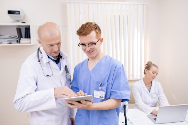 Dos médicos de sexo masculino en uniforme mirando la pantalla táctil mientras uno de ellos explica información médica o documento electrónico