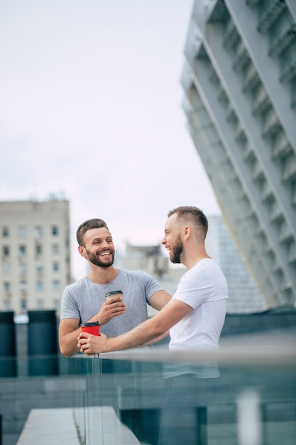 Dos mejores amigos barbudos jóvenes guapos en ropa casual están de pie al aire libre con tazas de café riendo y tienen una conversación.