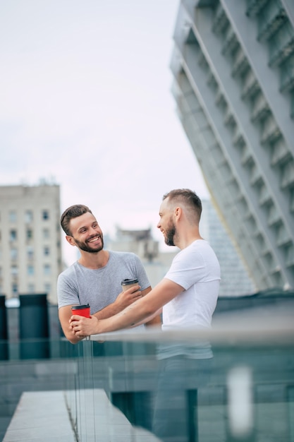 Dos mejores amigos barbudos jóvenes guapos en ropa casual están de pie al aire libre con tazas de café riendo y tienen una conversación.