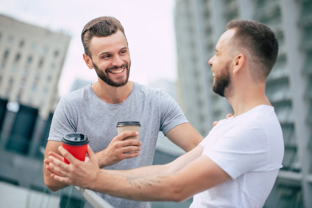 Dos mejores amigos barbudos jóvenes guapos en ropa casual están de pie al aire libre con tazas de café riendo y tienen una conversación.