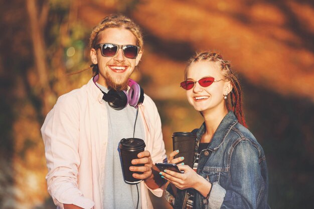 Dos mejores amigos hipster con rastas usando auriculares y gafas caminando juntos y bebiendo café en el soleado día de otoño