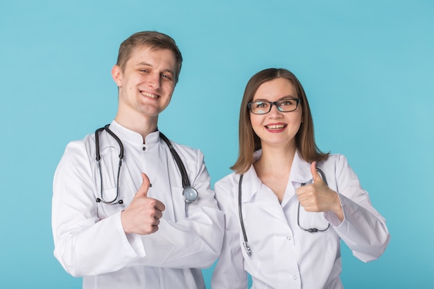 Dos mejores trabajadores médicos sonrientes profesionales inteligentes en batas blancas que muestran los pulgares para arriba de pie contra la pared azul.