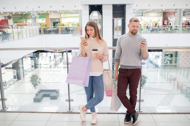 Dos millennials en ropa casual desplazándose en teléfonos inteligentes mientras están de pie junto a las barandillas dentro del centro comercial después de ir de compras