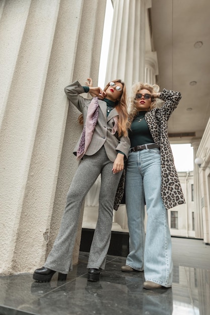 Dos modelos bonitas de moda chicas con gafas de sol frescas en ropa de otoño de moda con jeans abrigo de leopardo traje gris y zapatos se encuentran cerca de columnas antiguas en la calle