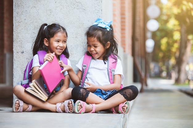 Dos muchachas asiáticas lindas del alumno que leen un libro juntos en la escuela con la diversión y la felicidad