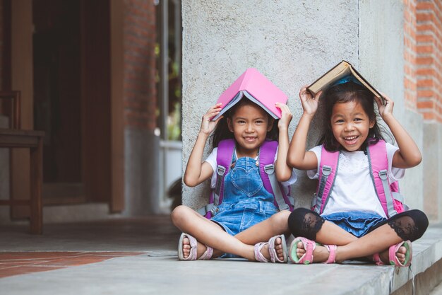 Dos muchachas asiáticas lindas del alumno que leen un libro juntos en la escuela con la diversión y la felicidad