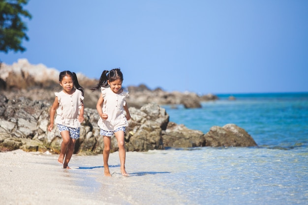 Dos muchachas asiáticas lindas del niño que se divierten jugar y correr en la playa juntas en vacaciones de verano