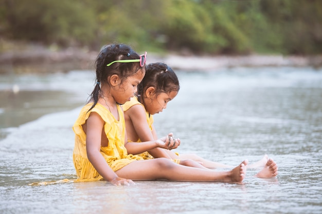 Dos muchachas asiáticas lindas del niño que se sientan y que juegan con la arena en la playa juntas