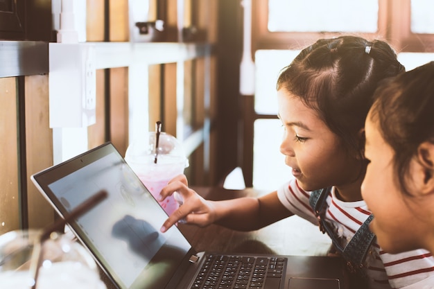 Foto dos muchachas asiáticas lindas del niño que usan y que juegan en el ordenador portátil en el café junto