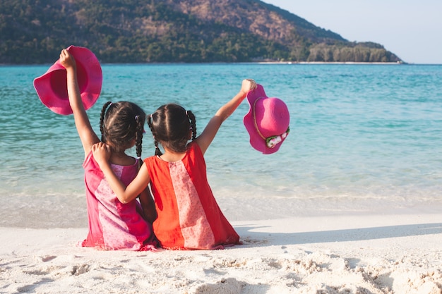 Dos muchachas asiáticas lindas del pequeño niño que sostienen el sombrero que se sienta y que juega junto en la playa