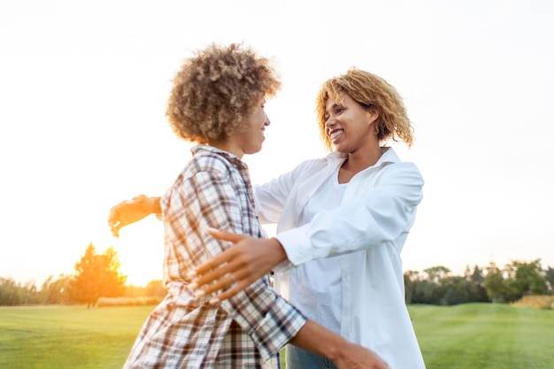 dos mujeres afroamericanas rizadas sonriendo y abrazándose en el parque en las hermanas del atardecer