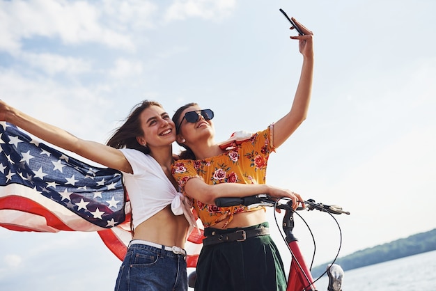 Dos mujeres alegres patrióticas con bicicleta y bandera de Estados Unidos en manos hacen selfie.