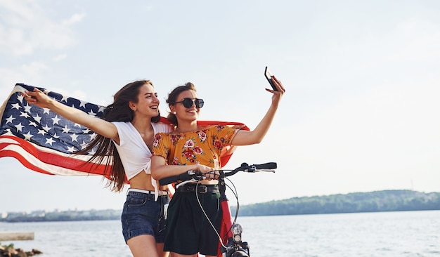 Dos mujeres alegres patrióticas con bicicleta y bandera de Estados Unidos en manos hacen selfie.