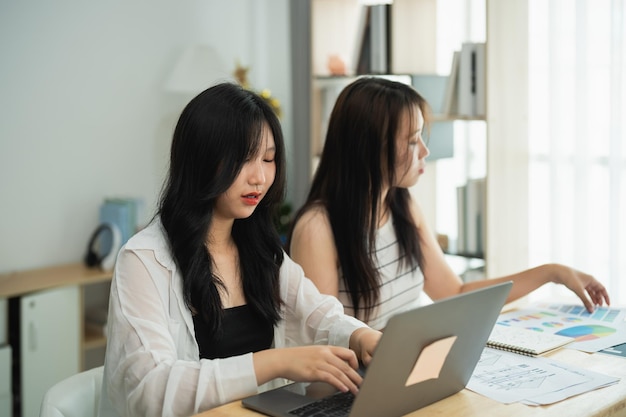 Dos mujeres asiáticas están sentadas en una mesa con computadoras portátiles una de ellas está escribiendo en una computadora portátil mientras la otra está mirando un pedazo de papel La escena está enfocada y seria