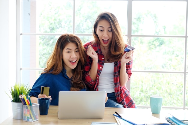 Dos mujeres asiáticas jóvenes que sostienen la tarjeta de crédito y usan una computadora portátil para hacer compras en línea