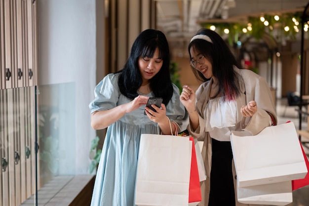 Dos mujeres asiáticas de pie juntas en un corredor de un centro comercial mirando un teléfono inteligente