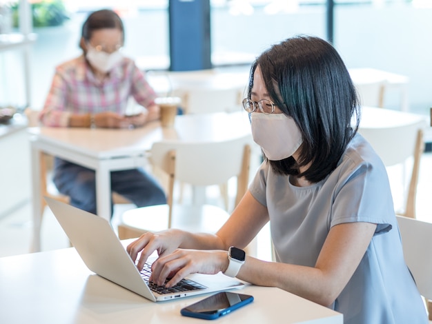 Foto dos mujeres asiáticas que usan mascarilla y usan teléfonos inteligentes y computadoras portátiles se sientan en mesas separadas para mantener el distanciamiento social de seguridad como un nuevo concepto de estilo de vida normal.