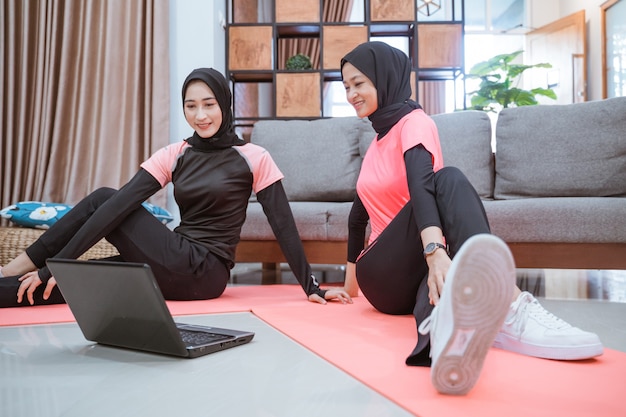 Dos mujeres asiáticas con ropa deportiva hijab se sientan en el suelo mientras se calientan las caderas juntas en la casa