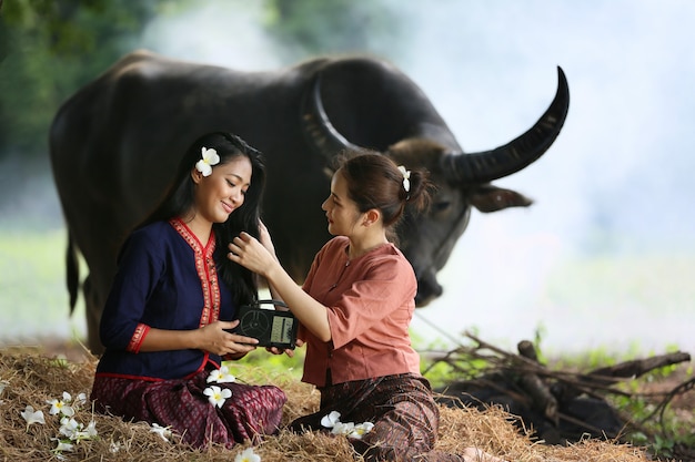 Dos mujeres asiáticas vistiendo trajes tradicionales tailandeses sentados en el campo, mientras escuchan la radio de estilo vintage junto a Buffalo