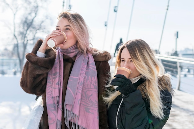 Dos mujeres bebiendo café de vasos de papel, de pie en el puente en invierno