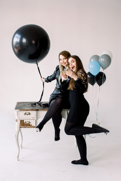 Dos mujeres bonitas jóvenes en vestidos negros que sostienen los globos de aire juntos, celebrando la despedida de soltera o cumpleaños cerca de la mesa blanca vintage con armarios, aislados en blanco