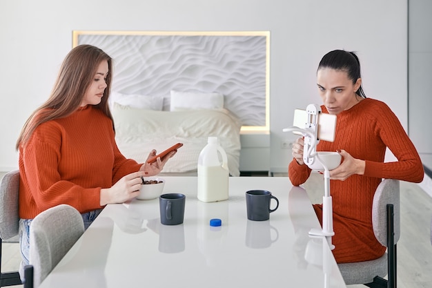 Dos mujeres desayunan en la mesa de la cena en la cocina y buscan información en sus teléfonos inteligentes.