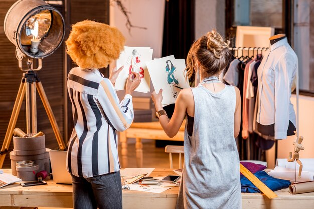 Foto dos mujeres diseñadoras de moda multiétnicas que trabajan con dibujos de ropa de pie en la hermosa oficina con diferentes herramientas y ropa de sastrería