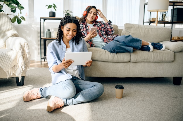 Dos mujeres disfrutan escuchando música en interiores. Bonitas novias en auriculares se relajan en la habitación, amantes del sonido descansando en el sofá, amigas ocios juntos