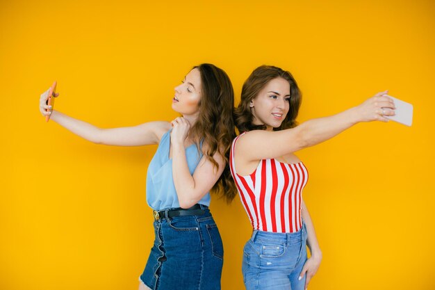 Dos mujeres elegantes felices en vestidos posando juntas y haciendo selfie en el teléfono inteligente