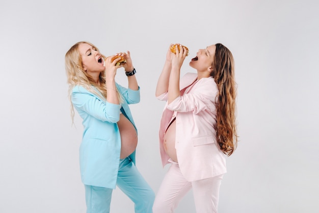 Dos mujeres embarazadas en trajes con hamburguesas en sus manos sobre un fondo gris.