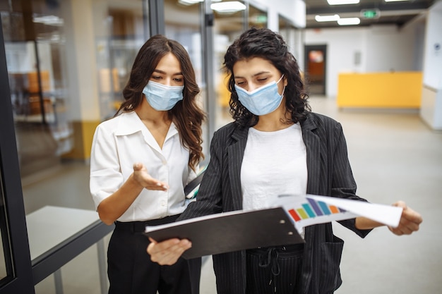 Dos mujeres empresarias discuten asuntos laborales en la oficina con máscaras médicas esterilizadas. Trabajando durante la cuarentena pandémica por coronavirus.