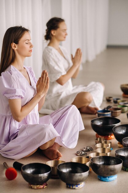 Dos mujeres están sentadas con cuencos tibetanos en posición de loto antes de una clase de yoga en el gimnasio.