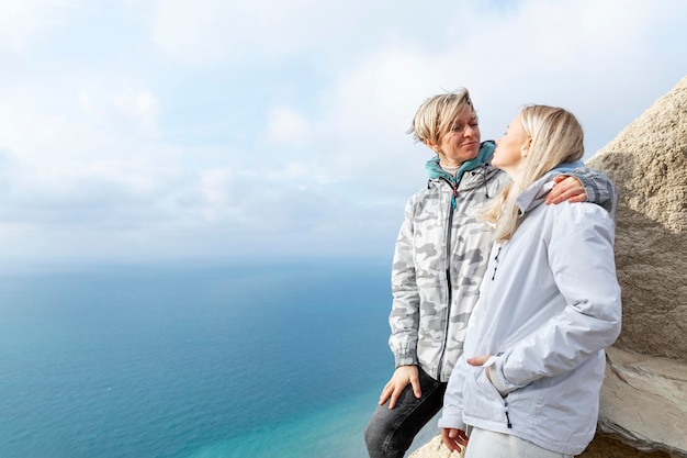 Foto dos mujeres felices en la cima de una montaña sobre el mar en un día soleado rubias en chaquetas deportivas abrazan y sonríen estilo de vida activo viajes y senderismo