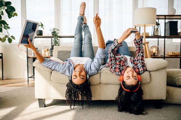 Dos mujeres felices disfrutan escuchando música al revés en casa. Bonitas novias en auriculares se relajan en la habitación, amantes del sonido descansando en el sofá, amigas ocios juntos