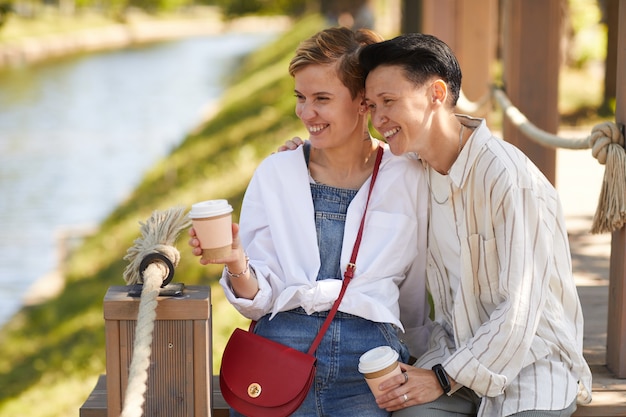 Dos mujeres felices tomando café y riendo mientras está sentado al aire libre en el parque