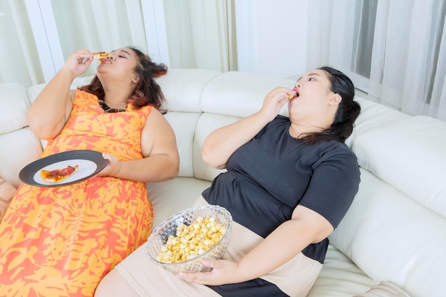 Dos mujeres gordas se ven llenas mientras comen en el sofá