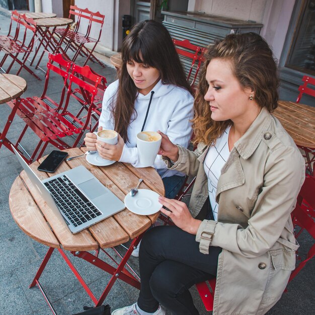 Foto dos mujeres jóvenes adultas bebiendo café y mirando una laptop en un café al aire libre