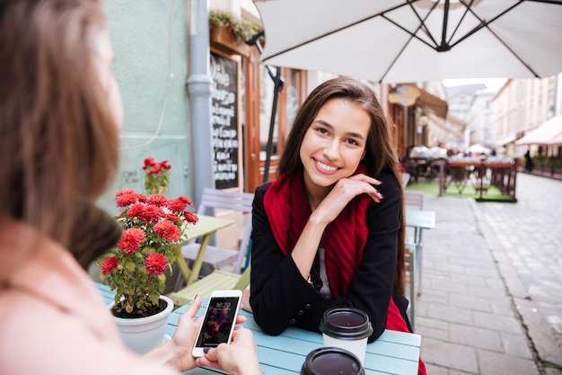 Dos mujeres jóvenes atractivas felices hablando y usando el teléfono celular en la cafetería al aire libre