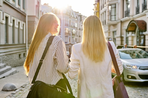 Dos mujeres jóvenes con bolsas para portátiles caminando por la calle de la ciudad al atardecer, vista trasera. Fondo de estilo urbano, estudiantes universitarias, colegas oficinistas mujeres