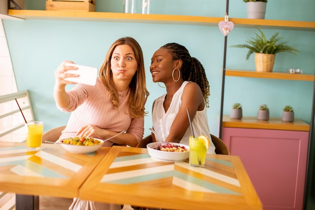 Dos mujeres jóvenes caucásicas y negras tomando selfie con teléfono móvil en el café