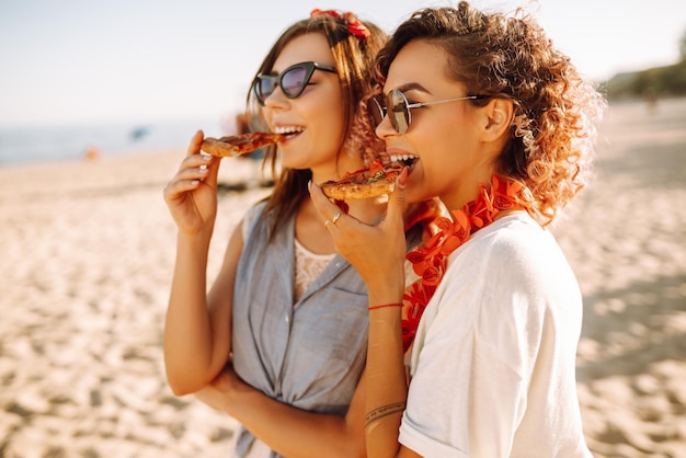 Dos mujeres jóvenes descansando juntas en la playa y comiendo pizza Concepto de comida rápida Vacaciones en la playa